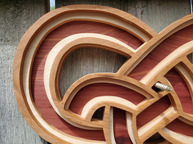 May include: A layered wooden wall art piece featuring an intricate, continuous knot design. The structure is composed of alternating light-colored wood and deep reddish-brown wood segments, creating a three-dimensional effect with a metal screw visible.