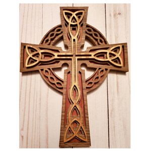 May include: A layered wooden Celtic cross featuring a dark brown outer frame, a reddish-brown middle layer, and a light tan top layer. The design includes intricate cut-out knotwork patterns and a circular ring intersecting the cross arms.
