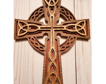 May include: A wooden Celtic cross with intricate knotwork design. The cross is made of two different shades of wood, creating a layered effect. The background is a light wood panel.