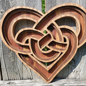 May include: A multi-layered wooden wall art piece shaped like a heart, featuring an intricate, interlocking Celtic knot design. The object is constructed from stacked wood panels with varying brown tones, creating a deep, dimensional effect.