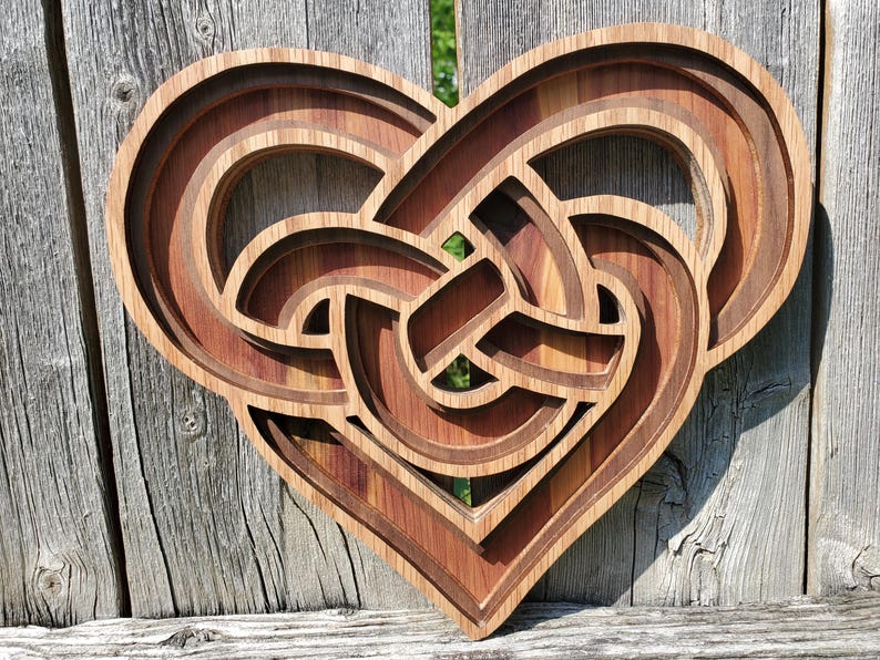 May include: A multi-layered wooden wall art piece shaped like a heart, featuring an intricate, interlocking Celtic knot design. The object is constructed from stacked wood panels with varying brown tones, creating a deep, dimensional effect.