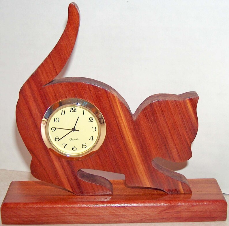 May include: A wooden cat clock with a light brown finish. The clock has a round face with black hands and black numbers. The clock is sitting on a wooden base.