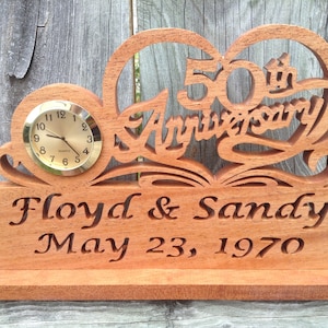 May include: A wooden desk clock featuring a cutout heart design with the text 50th Anniversary. The base is engraved with the names Floyd & Sandy and the date May 23, 1970. A small round clock face with a gold-toned dial is inset into the left side.