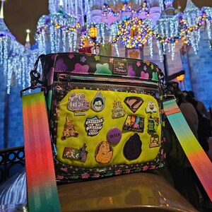 Peut inclure: Un sac bandoulière noir et violet avec une sangle arc-en-ciel, orné d'épingles et d'écussons. Le sac a une poche avant jaune néon avec diverses épingles. En arrière-plan, un château illuminé la nuit.