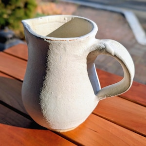 May include: A handmade, off-white ceramic pitcher with a curved spout and a large, rounded handle. The pitcher has a textured surface and sits on a wooden table, showcasing its rustic charm. The pitcher is ideal for serving beverages.