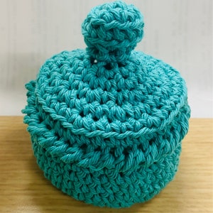 May include: A teal blue crocheted storage container with a round lid and a small pom-pom on top.