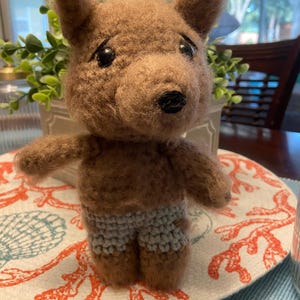 May include: A brown crocheted stuffed animal with a black nose and eyes. The animal is wearing blue and white crocheted shorts and is standing on a white and orange placemat.
