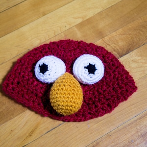 May include: A red crocheted hat with white and black eyes and an orange crocheted nose. The hat is a fun and whimsical accessory.