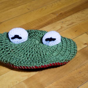May include: Green crocheted frog hat with white eyes and black moustaches. The hat has a red trim around the edge.