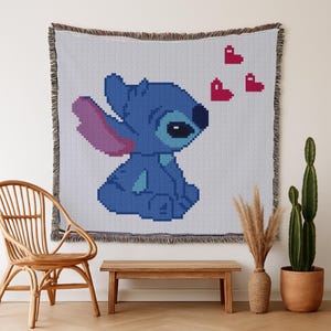 Op de afbeelding: Een geweven sprei met een gepixeld beeld van Stitch, in blauw met roze en rode accenten. De sprei heeft een gefranjerde rand. De sprei wordt aan een muur getoond.