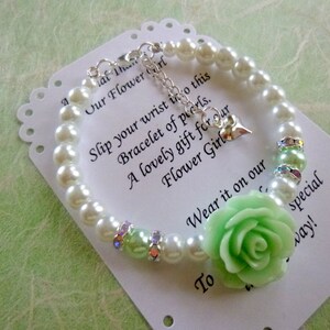 Op de afbeelding: Een witte parel armband met een lichtgroene roos bedel en een zilveren hart bedel. De armband ligt op een witte kaart met de tekst "Schuif je pols in deze armband van parels. Een mooi cadeau voor onze bruidsmeisje. Draag het op onze speciale dag!"