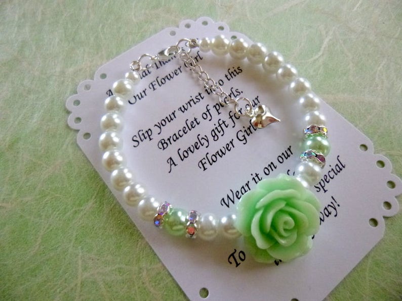 Op de afbeelding: Een witte parel armband met een lichtgroene roos bedel en een zilveren hart bedel. De armband ligt op een witte kaart met de tekst "Schuif je pols in deze armband van parels. Een mooi cadeau voor onze bruidsmeisje. Draag het op onze speciale dag!"