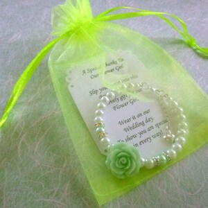 Op de afbeelding: Een groene organza tas met een witte kaart en een parel armband met een groene bloemenbedel. De kaart luidt: "Een speciale dank aan onze bloemenmeisje. Schuif je pols in dit mooie cadeau, onze bloemenmeisje. Draag het op onze trouwdag. Om te laten zien dat je op alle manieren speciaal bent!"
