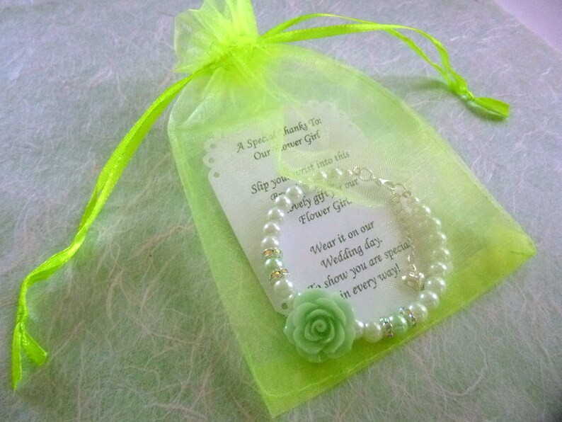 Op de afbeelding: Een groene organza tas met een witte kaart en een parel armband met een groene bloemenbedel. De kaart luidt: "Een speciale dank aan onze bloemenmeisje. Schuif je pols in dit mooie cadeau, onze bloemenmeisje. Draag het op onze trouwdag. Om te laten zien dat je op alle manieren speciaal bent!"
