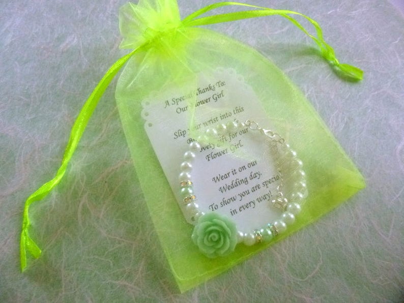 Op de afbeelding: Een groene en witte parel armband met een groene bloemenbedel wordt geleverd in een groene organza tas. De tas heeft een kaartje met de tekst "Een speciale dank aan onze bruidsmeisje. Schuif je pols in dit mooie cadeau voor onze bruidsmeisje. Draag het op onze trouwdag. Om te laten zien dat je op alle manieren speciaal bent!"
