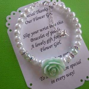 Op de afbeelding: Een witte parel armband met een lichtgroene bloemenbedel en een zilveren bedel. De armband ligt op een witte kaart met de tekst "Special Thanks To: Our Flower Girl. Slip your wrist into this bracelet of pearls. A lovely gift for Flower Girl. To s...re special in every way!"