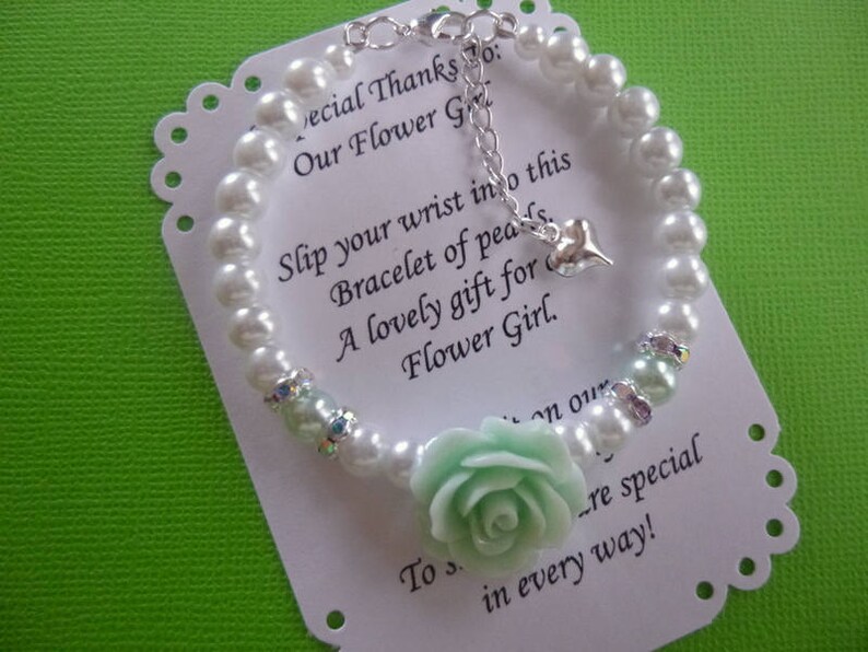 Op de afbeelding: Een witte parel armband met een lichtgroene bloemenbedel en een zilveren bedel. De armband ligt op een witte kaart met de tekst "Special Thanks To: Our Flower Girl. Slip your wrist into this bracelet of pearls. A lovely gift for Flower Girl. To s...re special in every way!"
