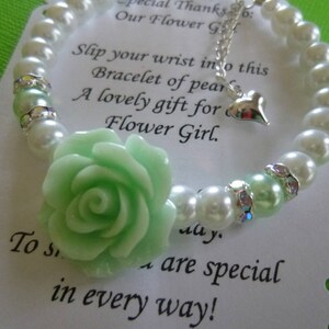 Op de afbeelding: Een witte parel armband met een lichtgroene bloem bedel en een zilveren bedel in de vorm van een hart. De armband ligt op een witte kaart met de tekst "Bedankt: Onze Bloemenmeisje. Schuif je pols in deze armband van parels. Een mooi cadeau voor een Bloemenmeisje. Voor iemand die op alle manieren speciaal is!"