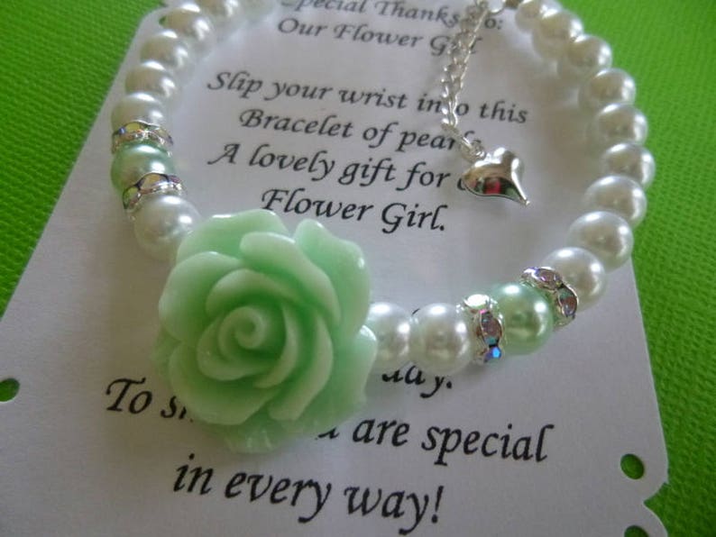 Op de afbeelding: Een witte parel armband met een lichtgroene bloem bedel en een zilveren bedel in de vorm van een hart. De armband ligt op een witte kaart met de tekst "Bedankt: Onze Bloemenmeisje. Schuif je pols in deze armband van parels. Een mooi cadeau voor een Bloemenmeisje. Voor iemand die op alle manieren speciaal is!"