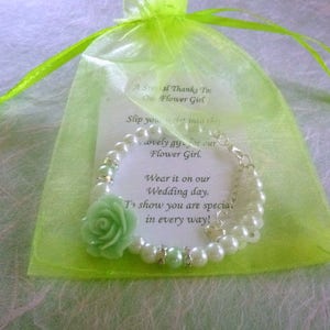 Op de afbeelding: Een groen en wit bloemenmeisjesarmband met een groene bloemenbedel en een kaart met de tekst "Een speciale dank aan onze bloemenmeisje. Trek deze mooie jurk aan voor onze bloemenmeisje. Draag het op onze trouwdag. Om te laten zien dat je op alle manieren speciaal bent!"