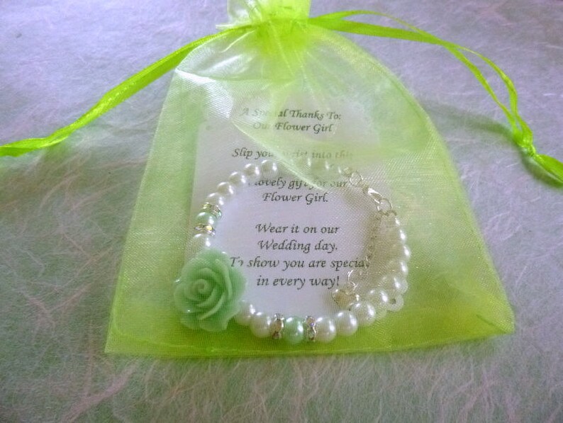 Op de afbeelding: Een groen en wit bloemenmeisjesarmband met een groene bloemenbedel en een kaart met de tekst "Een speciale dank aan onze bloemenmeisje. Trek deze mooie jurk aan voor onze bloemenmeisje. Draag het op onze trouwdag. Om te laten zien dat je op alle manieren speciaal bent!"