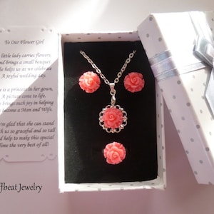 May include: A jewelry set featuring a necklace and earrings with coral-colored rose pendants, presented in a polka-dot gift box with a silver ribbon. The box contains a card with the text "To Our Flower Girl" and the brand name "Offbeat Jewelry".