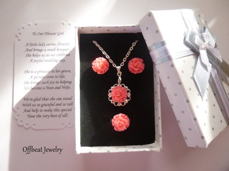 May include: A jewelry set featuring a necklace and earrings with coral-colored rose pendants, presented in a polka-dot gift box with a silver ribbon. The box contains a card with the text "To Our Flower Girl" and the brand name "Offbeat Jewelry".