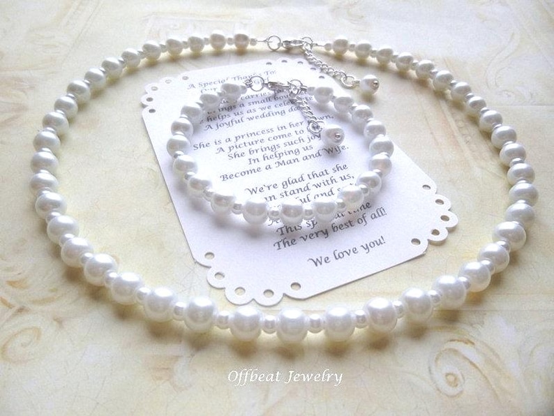 May include: A white pearl necklace and matching bracelet set. The jewelry is displayed on a white card with a scalloped edge and text. The set is likely for a wedding or special occasion. The jewelry is from Offbeat Jewelry.