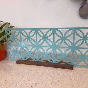 May include: A rectangular light blue metal screen featuring a repeating geometric pattern of intersecting circles. The screen is mounted vertically into a dark brown rectangular wooden base, providing a stable foundation for the decorative panel.