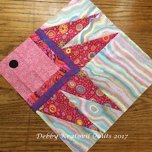 May include: A colorful quilt block with a geometric design featuring pink, red, blue, and yellow fabrics. The block is made up of triangles and squares, and the fabric has a variety of patterns, including polka dots, stripes, and swirls. The text "Debby Kratovil Quilts 2017" is visible in the bottom right corner of the block.
