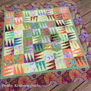 Op de afbeelding: Een kleurrijk quilt met een geometrisch patroon van driehoeken in tinten groen, oranje, paars en blauw. De quilt is omzoomd met een stoffen met een bloemenprint in tinten paars, oranje en bruin. De quilt is gesigneerd met "Debby Kratovil Quilts 2017".