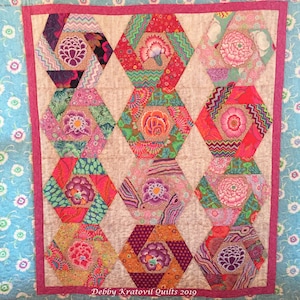 Könnte beinhalten: Ein farbenfroher Quilt mit floralem Muster. Der Quilt zeigt neun sechseckige Blöcke, jeder mit einem zentralen Blumenmotiv und umgeben von gemustertem Stoff in Rosa-, Rot-, Grün- und Lilatönen. Der Rand ist hellblau mit einem Blumenmuster.