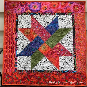May include: A vibrant, square quilt featuring a central star design. The star is composed of colorful, patterned fabrics in shades of red, blue, green, and orange, set against a white background with black polka dots. The quilt has a floral border and the text "Debby Kratovil Quilts 2017".