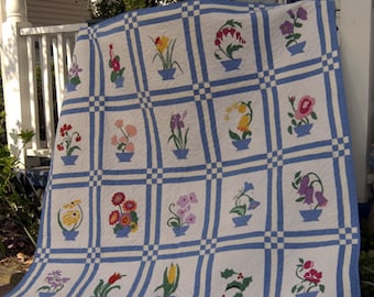 Könnte beinhalten: Eine weiße Bettdecke mit einem blau-weißen Karomuster. Jedes Quadrat zeigt eine andere gestickte Blume in einem blauen Topf.