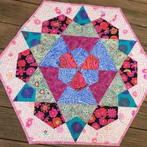 May include: A pink and white octagonal table runner with a colorful geometric pattern in the center. The pattern features pink, blue, green, and black fabrics with floral and abstract designs.