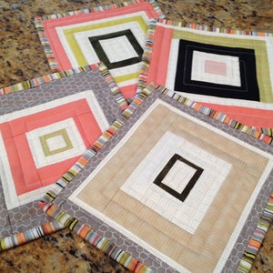 May include: Four square-shaped quilted fabric coasters with different colored borders and inner squares. The coasters are made of a light beige fabric with a subtle texture. The borders are made of a variety of colors, including gray, orange, yellow, and green.