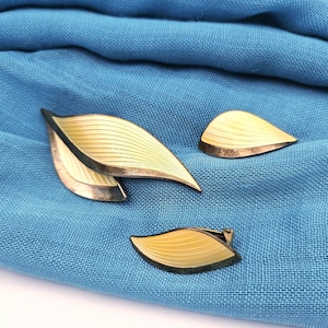 Peut inclure: Un ensemble de bijoux vintage en forme de feuilles. L'ensemble comprend une grande broche et deux petites pièces, toutes avec un design en émail jaune pâle avec une bordure en métal argenté. Les bijoux sont présentés sur un fond en tissu bleu.
