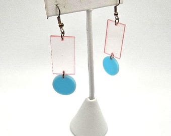 Op de afbeelding: Een paar oorhangers met een roze rechthoekige vorm en een lichtblauwe cirkel die aan elke oorhanger hangt.