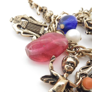 May include: A gold chain necklace with multiple charms, including a pink glass bead, a blue glass bead, a white pearl, a gold cherub, and a gold lyre.