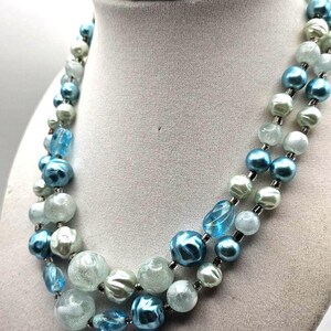 May include: A two-strand necklace with blue, light blue, and white beads. The beads vary in size and shape, with some being round and others more oblong. The necklace is displayed on a gray jewelry bust.