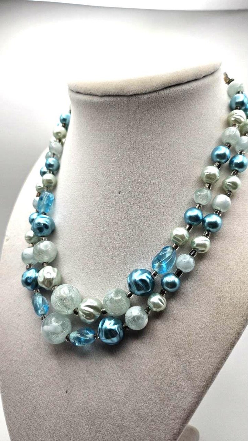 May include: A two-strand necklace with blue, light blue, and white beads. The beads vary in size and shape, with some being round and others more oblong. The necklace is displayed on a gray jewelry bust.
