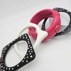 May include: A collection of colorful bangles in various shapes. The set includes a black square bangle with white polka dots, a white geometric bangle, a bright pink round bangle with black polka dots, and a black and white speckled round bangle.