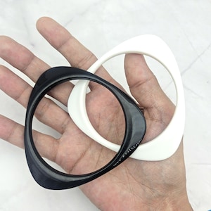 Puede incluir: Dos brazaletes geométricos en una mano. Uno es negro, el otro blanco. Cada brazalete tiene una forma triangular redondeada. Los brazaletes son de material brillante, probablemente joyería.