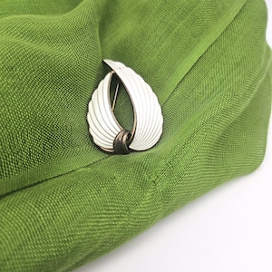Peut inclure: Broche vintage blanche et dorée avec un motif d'aile stylisé. La broche est posée sur un tissu vert texturé, créant un contraste saisissant. La broche présente un design incurvé et ouvert avec un accent central.
