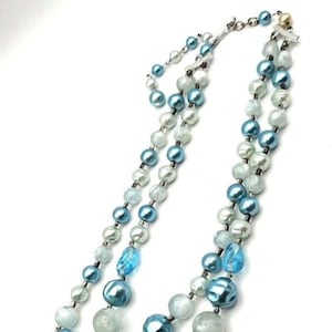 May include: A layered necklace featuring alternating blue, light blue, and white beads. The necklace has a silver clasp and is displayed against a white background. The beads vary in size and texture, creating a visually appealing accessory.