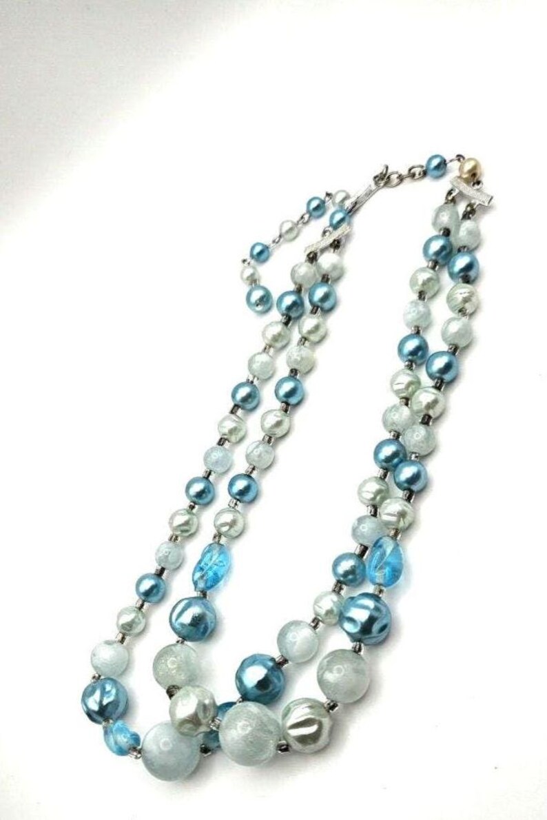 May include: A layered necklace featuring alternating blue, light blue, and white beads. The necklace has a silver clasp and is displayed against a white background. The beads vary in size and texture, creating a visually appealing accessory.