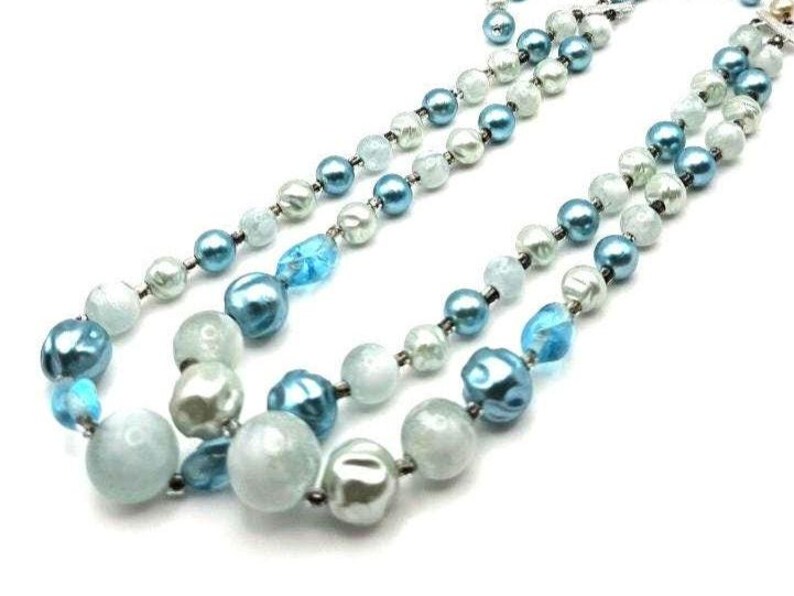 May include: A multi-strand necklace featuring a mix of blue, teal, and white beads. The beads vary in size and shape, including round and irregular forms. The necklace is displayed against a white background, highlighting the colors and textures of the jewelry.