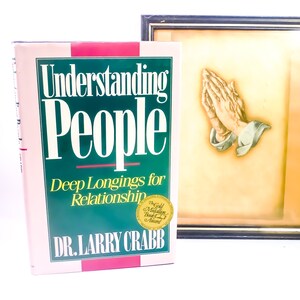May include: A pink and green hardcover book titled "Understanding People: Deep Longings for Relationship" by Dr. Larry Crabb. The book has a gold sticker that says "The Gold Medallion Book Award". The book is next to a framed picture of two hands clasped in prayer.