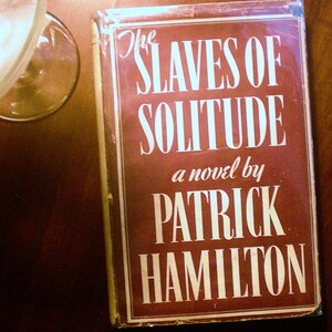 May include: A red book with the title "The Slaves of Solitude" and the author "Patrick Hamilton" printed in white.