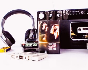 May include: A black cassette player with a silver dial and buttons. There are several cassette tapes stacked next to it, including tapes by Motley Crue, Cheap Trick, Billy Squier, and Jim Hendrix. There are also headphones and a cassette tape on the table.